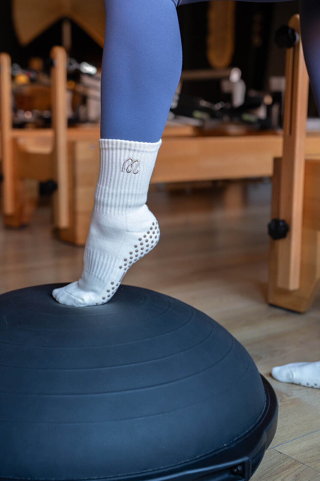 Gummi Socks white crew-length grip socks with cushioned sole and grey grip dots, worn in a Pilates studio on a balance dome, suitable for reformer, yoga and barre.