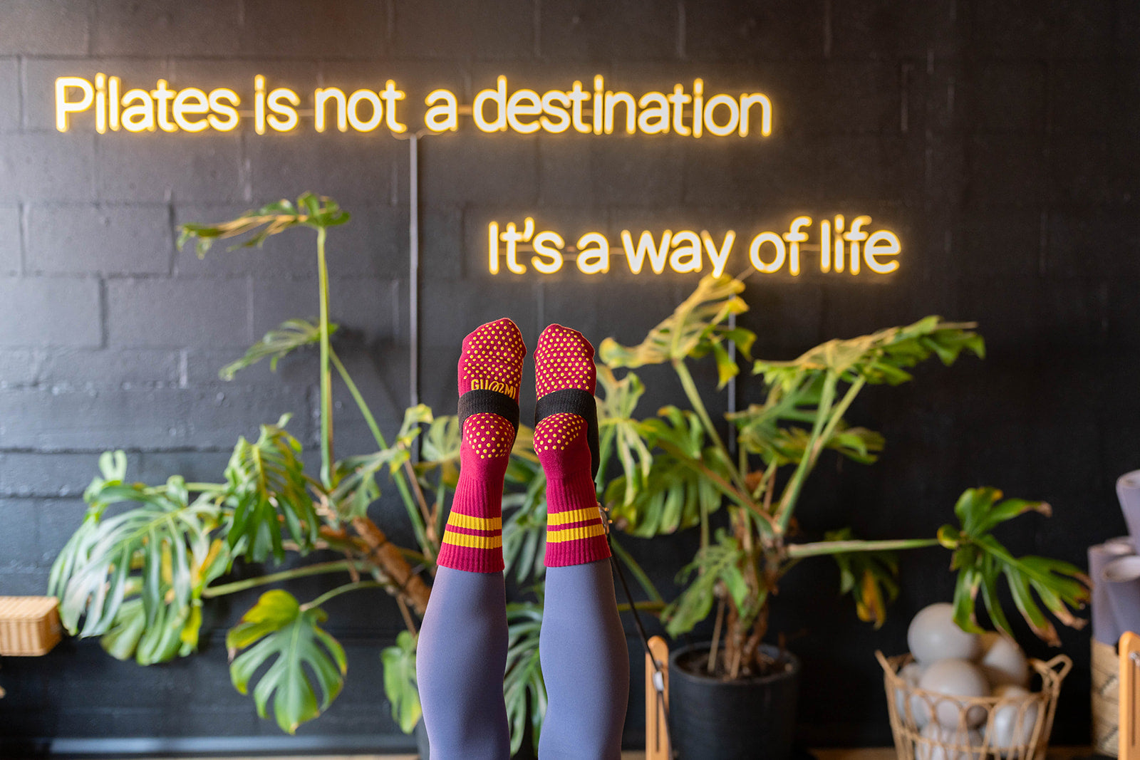 Person wearing colorful socks infront of text 'Pilates is not a destination, it's a way of life' on a dark wall with plants.